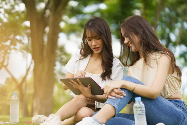 Couple Asian woman relaxing together in the park in natural day light while working with tablet to explore social network. Concept lifestyle smart working anywhere.