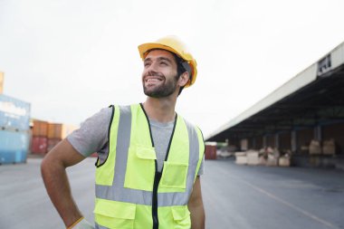 Arka planda konteynır avlusunun önünde duran, güvenlik yeleği ve başlık takmış gülümseyen beyaz adam portresi. Bölgede çalışan iyimser adam.