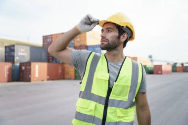 Arka planda konteynır avlusunun önünde duran, güvenlik yeleği ve başlık takmış gülümseyen beyaz adam portresi. Bölgede çalışan iyimser adam.