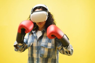 VR gözlüklü ve boks eldivenli genç bir kadının portresi. Gardiyan pozisyonunda el ele tutuşuyor, boks maçlarını seviyor, oyuncu, sarı arka planda hobi ediniyor.. 