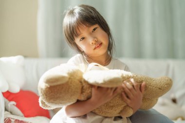 Asyalı tatlı kızın portresi oyuncak ayıya sarılıyor. Kameraya gülümsüyor. Evinde mutlu ve sakin hissediyor..