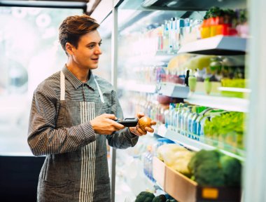 Marketteki taze meyve dükkanında gizli hayat tarzı genç bir adam raftaki ürünlerin kalitesini kontrol ediyor. Maskesiz genç bir erkek, mini sanatlarda çalışıyor..