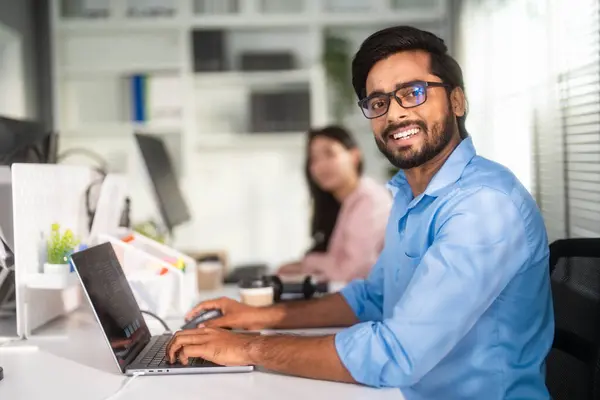 Portrait of an Indian man smiling while working with a laptop at the office and looking at the camera.