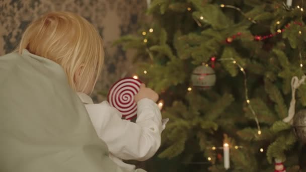 金发小女孩站在床和圣诞树之间的卧室里 手里拿着糖果 手指在一个圆圈里移动着 慢动作高质量的4K镜头 — 图库视频影像