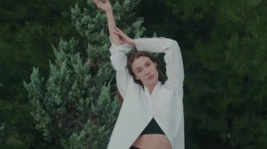Çekici genç bir kadın, ellerini yeşil bitkilerin arka planında kaldırıyor, sonra ellerini indiriyor ve yana dönüyor. Sürekli kameraya bakıyorsun. Beyaz moda gömleği. Yavaş çekim.
