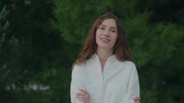 Parktaki yeşil bitkilerin arka planında gülümseyen bir kadının portresi. Uzun kıvırcık esmer saçlı kız. Genç bir bayanın yakın çekimi. Özgürlük ve mutluluk hissi. Yavaş çekim.