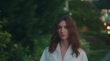 Şık, kendine güvenen genç bir kadın, akşam sokakta yalnız başına yürürken kameraya bakıyor. Gülümsüyor, saçlarına dokunuyor ve şehir ışıklarının önünde dönüyor. Moda tarzı. 4k