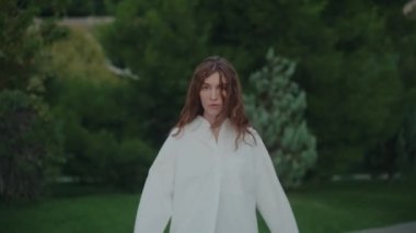 Beyaz gömlekli genç bir kız yeşil bitki ağaçlarının arka planında kameraya bakarak şehir parkında yürüyor. Kendine güvenen ciddi moda tarzı. Yavaş çekim.
