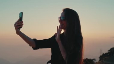 Dişi gezgin, gün batımında akıllı telefon görüşmesiyle dağ manzaralı bir yerde duruyor. Mutlu İzlenimler Tatili Genç kadın güneş gözlükleri uzun esmer saçlar.