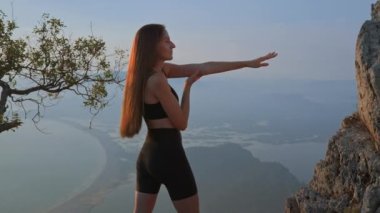 Beyaz tenli, ince, güzel bir kadın gün batımında kayaların arasında duruyor ve kollarını farklı yönlere açıyor. Sahildeki deniz adalarında güzel bir atletik kadının arkasından bakın..