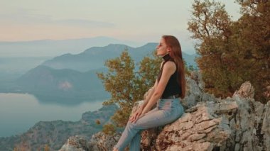 Güneş gözlüğü takan mutlu genç bayan gezgin dağın tepesindeki kayada oturur ve inanılmaz güneşin batışının tadını çıkarır. Özgürlük Adası Halkı, Mutluluk Kavramı. Yüksek kalite 4k görüntü