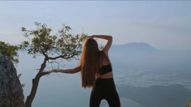 Arkaya bakan ince bir kadın, okyanusa ve adalara tepeden bakan kayaların arasında duruyor. Kolları farklı yönlere açılmış. Güneşli bir günde arkadan bakan kadın dışarıda spor yapıyor..