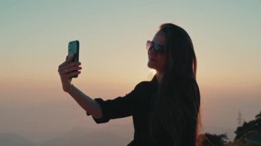 Yakın plan siluet, uzun saçlı, güneş gözlüklü genç kız elinde akıllı telefon tutan blogcu sosyal medya için video yapıyor. Genç, çekici, beyaz bir kadın. Sinematik 4k görüntü