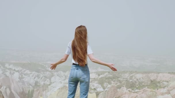 后视镜身穿休闲装 一头长长的黑发的兴奋的女徒步旅行者高举双手站在山顶上 享受着寂静和温暖的微风 卡帕多西亚 慢镜头 — 图库视频影像