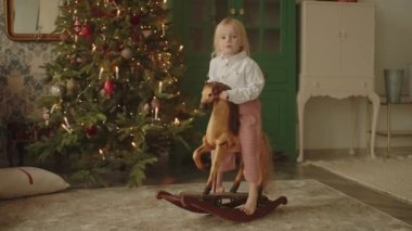 Küçük sarışın kız Noel arifesinde oyuncak atın üstünde yalnız başına oturuyor. Çocuk 3-4 yaşlarında, hediyeler hayal ediyor, ebeveynler mutfakta meşgulken misafirleri bekliyor. Fransız tarzı dekorasyonlar. 4K.