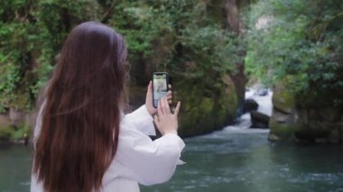 Genç bayan gezgin, şık görünümlü, akıllı telefonuyla bir dağ gölünün fotoğrafını çekiyor. Beyaz ırktan çekici bir genç kadın, esmer, uzun saçlı..