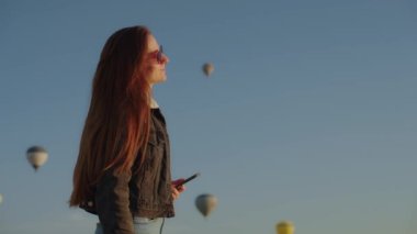Genç ve güzel bir kadın, gün doğumunda güneşin tadını çıkarıyor ve sıcak hava balonları uçuşuyor. Arka planda açık mavi gökyüzü var. Kapadokya Sıcak hava balonu festivali 4K ağır çekim.