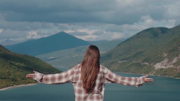 自由的概念 女徒步旅行者 举着胳膊 享受着温暖的微风和高山湖景 后视镜 年轻的白人女人 长长的黑发 时髦的衬衫 — 图库视频影像