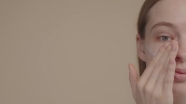 Portrait of half a yoang woman face in close up, on light background, looking into the camera, applying facial nourishing cream. Enjoy process while taking care perfect skin. Slow motion 4K