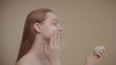Happy attractive young woman applying moisturizer Side view Young lady with long blonde hair rubbing the cream on her cheek, enjoying the natural beauty and healthy clean soft well-groomed skin SLO MO