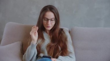 Smiling young woman customer holding glasses credit card and smartphone sitting on couch at home. Happy female shopper using instant easy mobile payments making purchase in online store. E-banking app