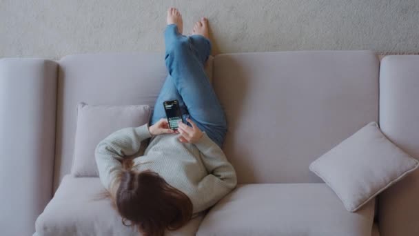 スマートフォンを持っている若い女性 モバイルアプリを使用して ソファに座っている間に計画を書いて 女性のチャット 自宅でソファの上にラウンジながら ソーシャルメディアチャット トップビュー — ストック動画