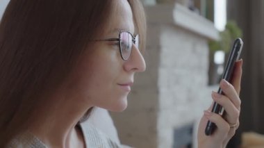 4K Close-up, profile view, face of a woman with vision glasses, holding a smartphone, searching the Internet, using app, touching screen, zooming in on the gadget screen with her fingers. Slow motion.