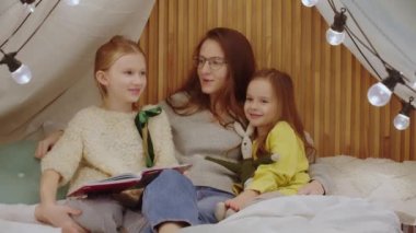 Anne ve iki kız yatak odasında oturmuş kitap okuyor ve eğleniyorlar. Büyük kız okuma yeteneğini gösteriyor. Bebek bakıcısı çocuklarla eğleniyor. 4k