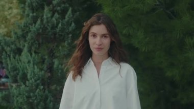 Large static shot of a cheerful and beautiful young college student with a smile on her face looking at the camera in broad daylight, green trees in the background. Slow motion footage. 4k