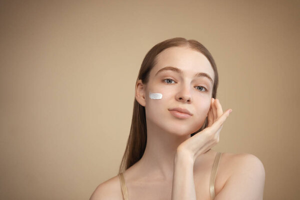 Facial skin care, applying moisturizing cream. Beautiful Caucasian young lady with blue eyes on an isolated background with a strip of face cream on her check, looking into the lens. 