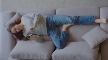 Mutlu, rahat bir kız cep telefonuyla mobil uygulamalar kullanıyor kanepeye uzanmış komik videolar izliyor, tembel genç bir kadın sosyal medyada sohbet ederken evde otururken sosyal medyada eğleniyor.