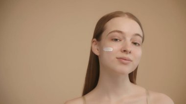 Sarışın ve mavi gözlü genç beyaz kız, izole edilmiş bir ışık arka planıyla ortaya çıkan yüz kreminin nemlendirici faydalarını sergiliyor.