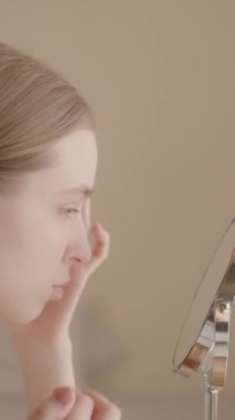 Endişeli genç kadın aynaya bakar, cildini inceler. Deri durumundan memnun olmayan mutsuz Z jenerasyonu kadın, profesyonel cilt bakım ürünlerini düşünüyor. Dikey video