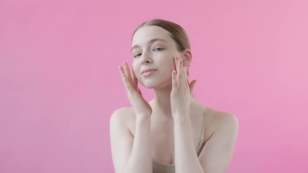 健康的で穏やかな女性は カメラを見て 孤立したピンクの背景に滑らかな肌で顔に触れています スキンケア広告 アンチにきび製品 皮膚クレンザー 保湿剤 フェイスリフト スローモーション — ストック動画