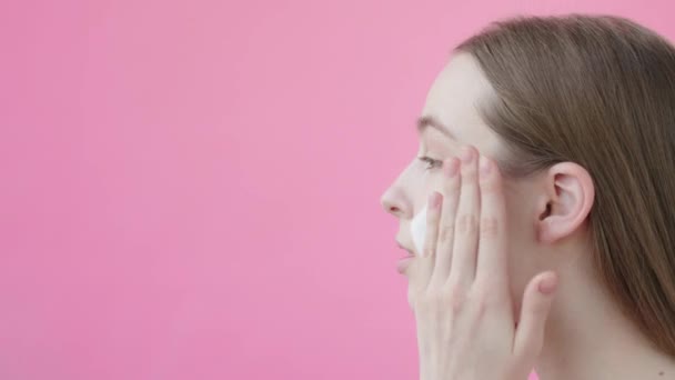 若い女性は顔泡で顔を洗う サイドビュー ピンクの背景 手の動き クレンジング頬スタジオショット にきび対策広告コンセプトスキンケアルーチン ピンクの背景美容コレクション — ストック動画