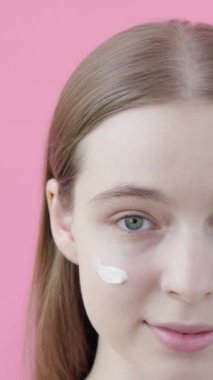 Yakından güzel bir portre genç sarışın beyaz kadın yanağına güneş kremi sürmüş, kameraya bakıyor ve gülümsüyor. Güneş kremi. Stüdyo çekimi. Pembe arka plan güzellik koleksiyonu. Dikey video