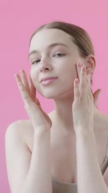 Genç, sağlıklı, sakin bir bayan kameraya bakıyor, izole edilmiş pembe arka planda pürüzsüz bir cilde dokunuyor. Cilt bakımı reklamı, sivilce önleyici ürünler, serum, nemlendirici, yüz gerdirme. Ağır çekim. Dikey video
