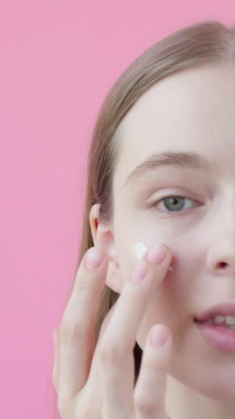 若いブロンドの女性は頬に保湿剤を適用します 顔のスキンケアの概念 ピンクの背景美容コレクション 垂直ビデオ — ストック動画