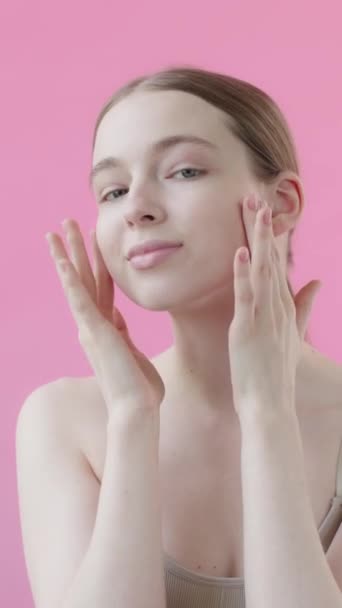 健康的で穏やかな女性は カメラを見て 孤立したピンクの背景に滑らかな肌で顔に触れています スキンケア広告 抗にきび製品 保湿剤 フェイスリフト スローモーション 垂直ビデオ — ストック動画