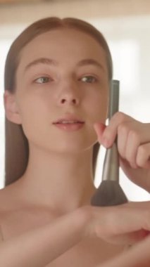 Yansımanın yakın çekimi, beyaz tenli genç bir kadın elinde fırçayla yanaklarına allık tozu sürüyor, makyaj yapıyor, sakin, kendine güvenen güzel bir bayan banyoda makyaj yapıyor, dikey video çekiyor. 