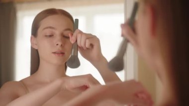 Yansımanın yakın çekimi, beyaz tenli genç bir kadın elinde fırçayla yanaklarına allık tozu sürüyor, makyaj yapıyor, sakin, kendine güvenen güzel bir bayan sabah makyajı yapıyor, yavaş çekim. 