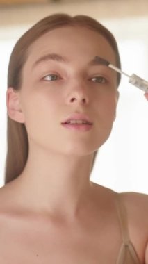 Makyaj. Kendine güvenen beyaz bir kadın kaşlarını kozmetik fırçayla fırçalıyor, günün başlangıcından keyif alıyor. Kaş şekillendirme, makyaj konsepti. Dikey video