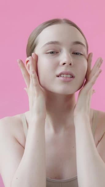 20代の若い女性は 顔に血清を適用し カメラを見て ピンクの背景 顔の美容製品を使用した完璧なきれいな肌の魅力的なプロのモデル コンセプトスキンケア広告 バーティカルビデオ — ストック動画