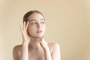 Bej arka planda genç, sağlıklı, sakin bir kadın pürüzsüz tenli, cilt bakımlı bir yüze dokunuyor. Anti-sivilce ürünü, serum, temizleyici, nemlendirici, yüz gerdirme. Güzel bir genç bayanın portresi.