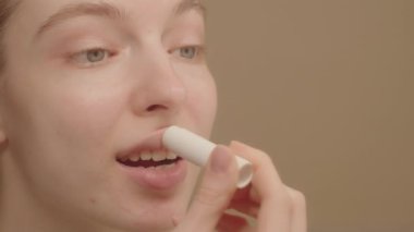 Beyaz tenli çekici bir kadının dudak kremi sürerken ve nemlendirici etkinin keyfini çıkarırken üç çeyrek portresi. Stüdyo çekimi bej arka plan Boş alan dudak kremi reklam konsepti.