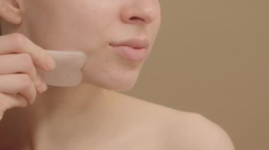 Gua sha. Yüz masajı. Genç bir kadın kuvars taşı kullanıyor. Rahatlamış genç bir kadın gülümsüyor ve hareketlerden zevk alıyor yüzüne bir gua sha taşıyla dokunuyor..