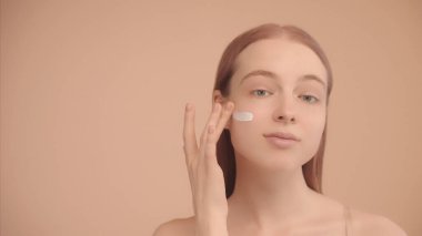 Yanağında yalıtılmış şeftali arkaplan kremi lekesi olan beyaz bir kadının portresi. Kadın nemlendirici krem sürüyor, kameraya bakıyor..