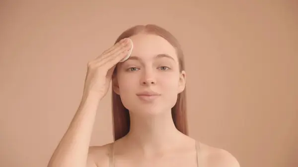 Woman uses a cotton pad to clean her face. Portrait of a young woman on an isolated peach background, looking at camera. The concept of makeup removal or application of exfoliant.