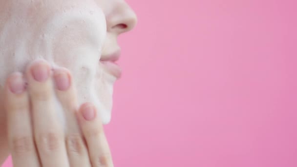 年轻女孩用面部泡沫洗脸 工作室拍摄 年轻女士在孤立的粉红色背景应用清洁剂泡沫 日常护肤广告的概念 慢动作 — 图库视频影像