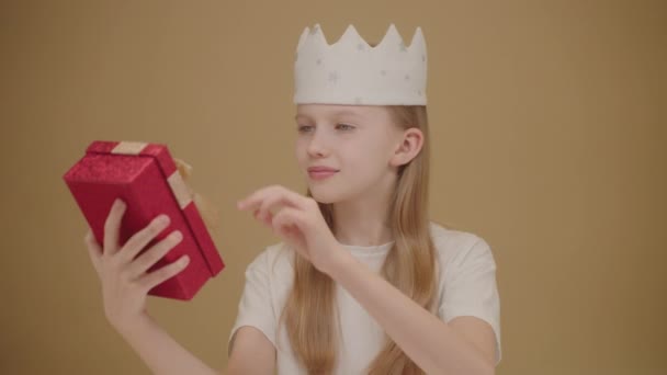 彼女の頭の上に冠を持つかわいい十代の少女は 贈り物を受け取りました 驚いた子供 誕生日ギフトボックス 幸せな子供 ベージュの背景で孤立 幸せな誕生日 — ストック動画